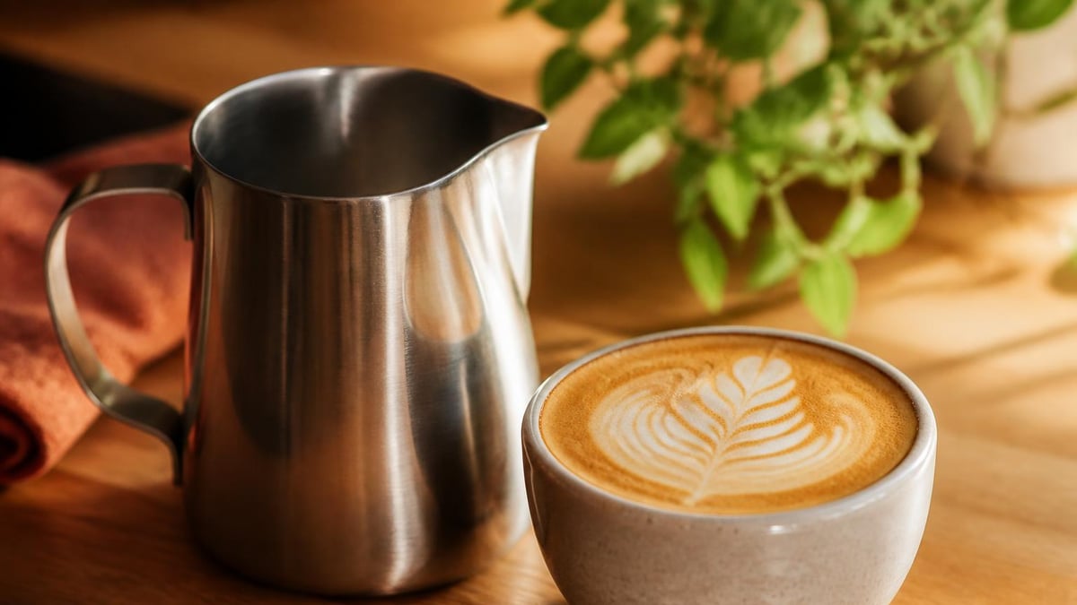 A stainless steel milk pitcher with a tapered spout sitting beside a latte on a wooden counter, warm natural light